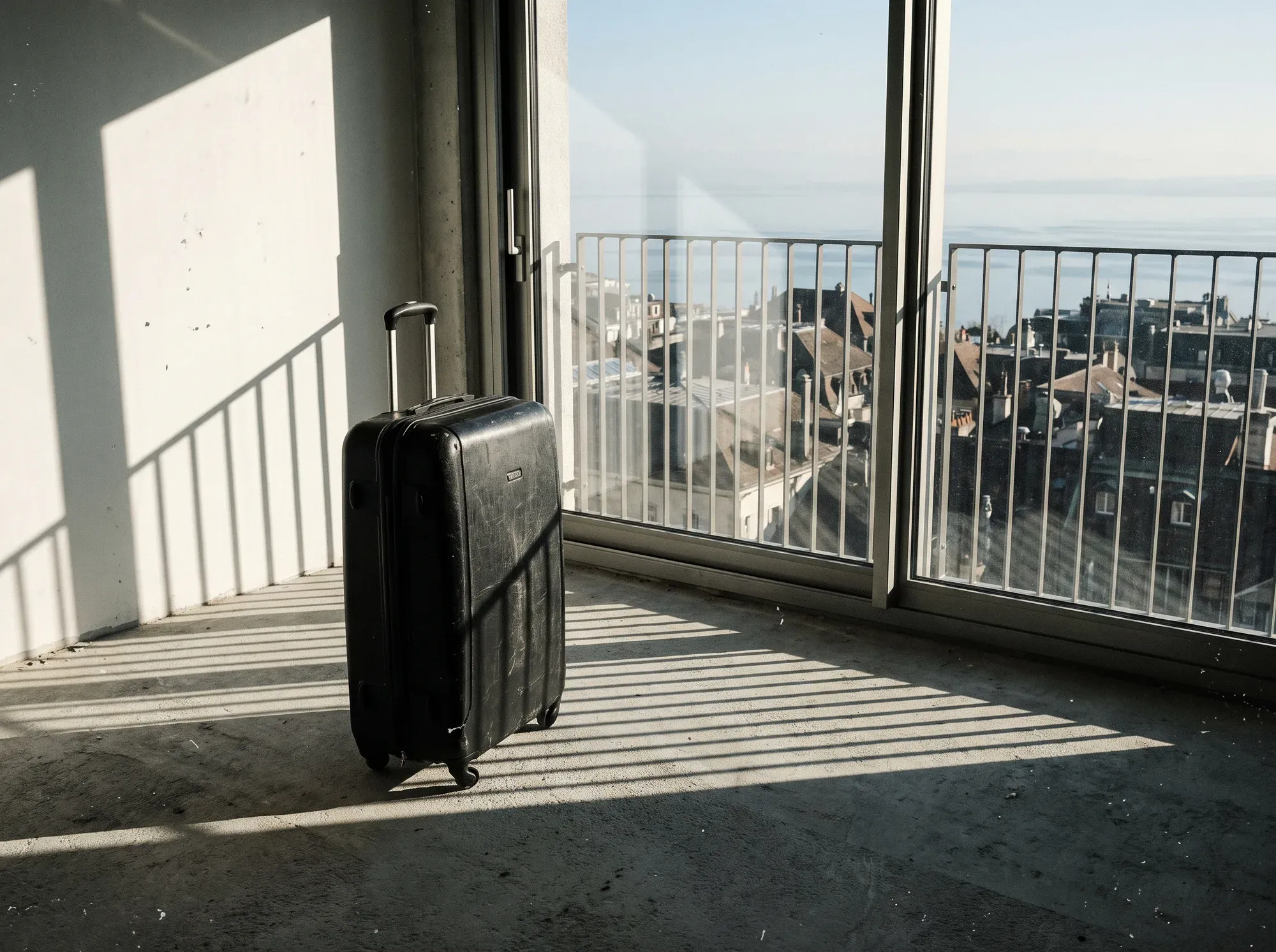 Valise noire dans un appartement vide en béton, toits de Lausanne et Léman derrière la vitre.