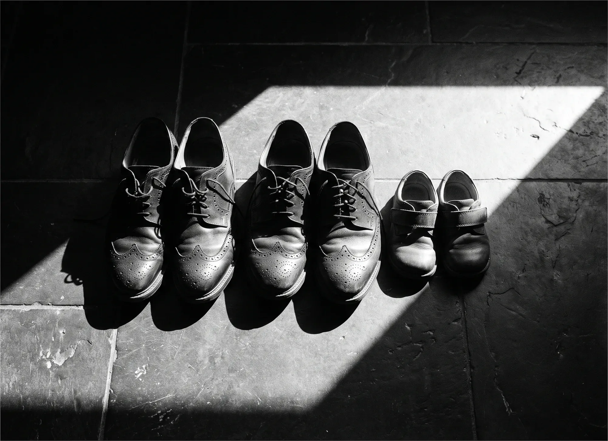 Chaussures de deux adultes et d’un enfant alignées sur un sol de pierre sombre.